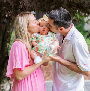 Parents sharing kisses with their baby wearing a floral outfit under dark foliage.