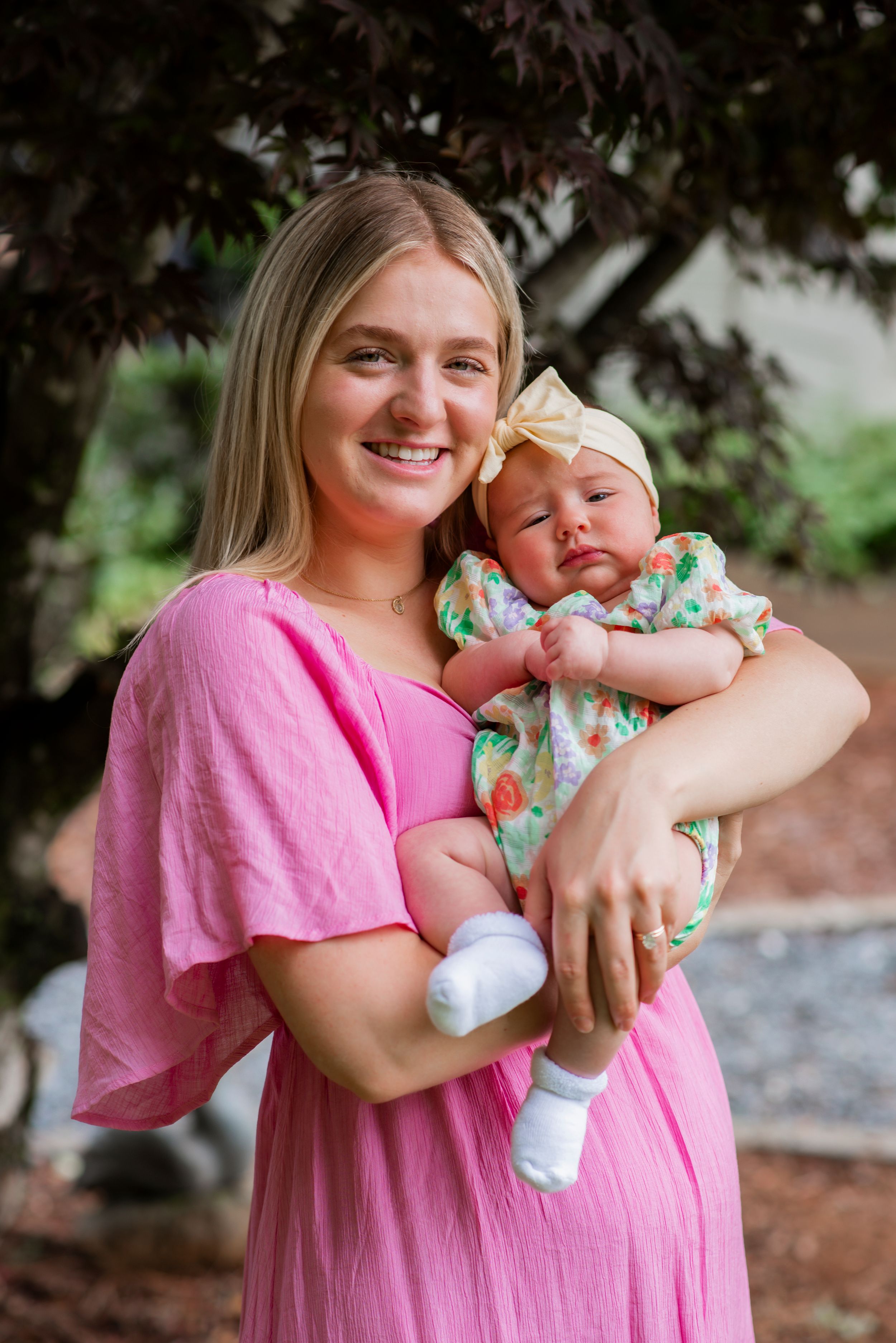Woman in pink dress holds sleeping infant wearing floral outfit outdoors.