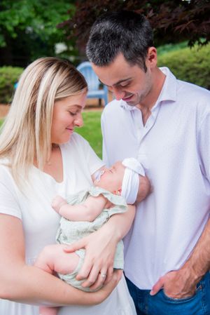 Parents in white shirts admiring their sleeping newborn baby outdoors.