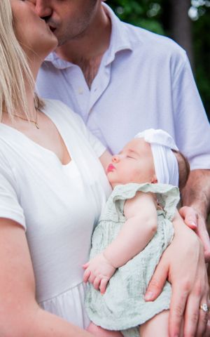 A sleeping baby wearing a white bow headband is held by parents in white shirts outdoors.