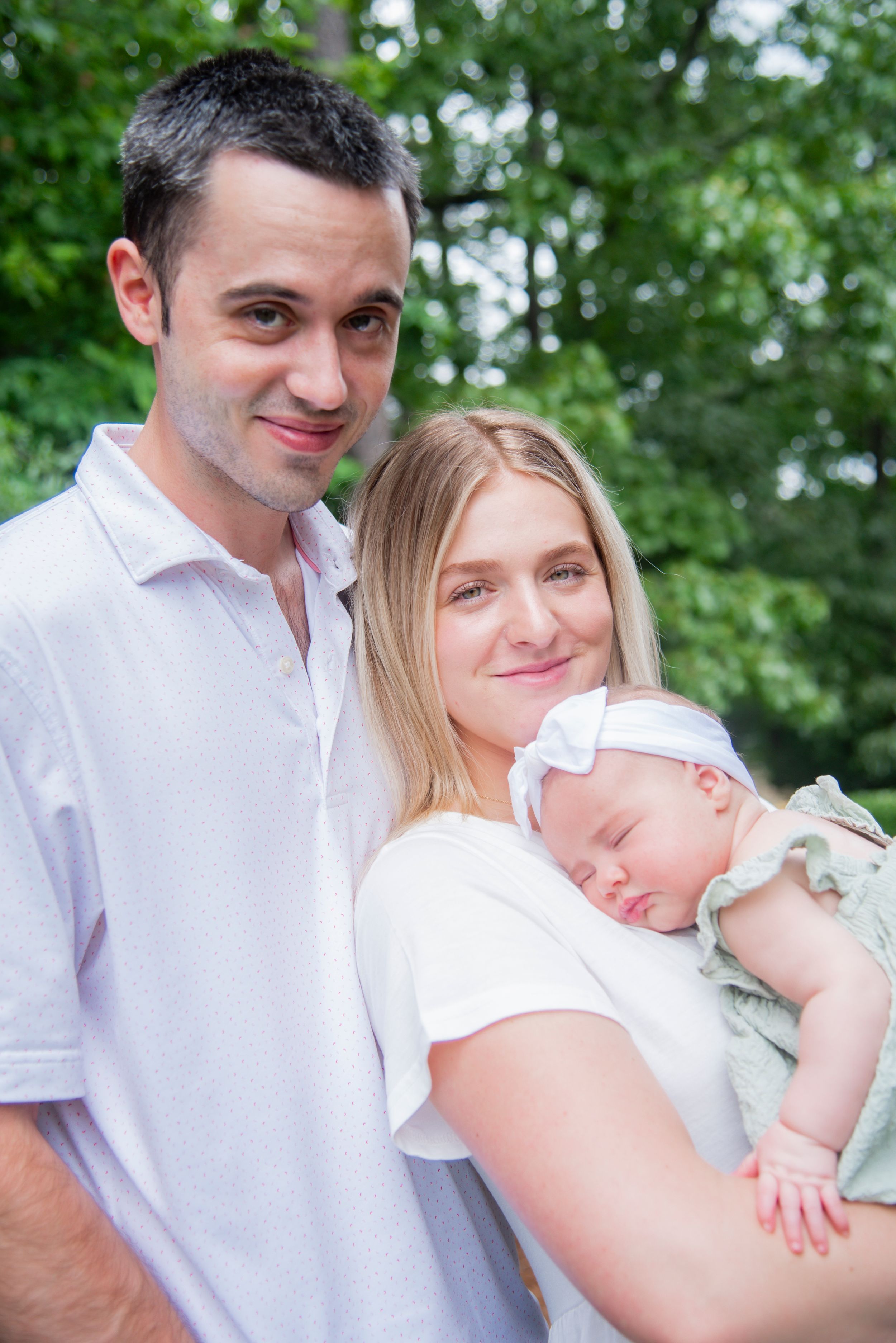 A family portrait outdoors with a sleeping baby wearing a white bow and mint green outfit.
