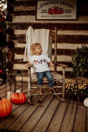 Rustic fall scene with toddler in rocking chair by log cabin wall with pumpkins and autumn decor.