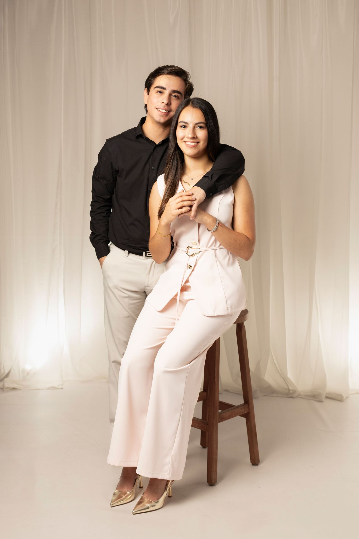 Young couple posing in studio, man in black shirt, woman in pink jumpsuit seated on wooden stool, white curtain backdrop.