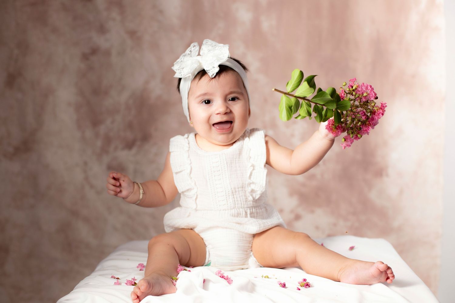 Baby in white dress sits on pink blanket while playing with fresh flowers in a heartwarming photography session.