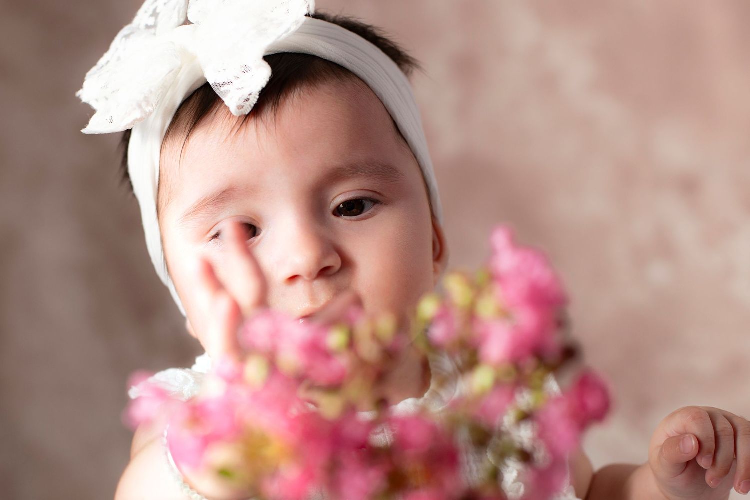 Baby wearing a white headband poses with pink flowers in the foreground during a professional photo session.