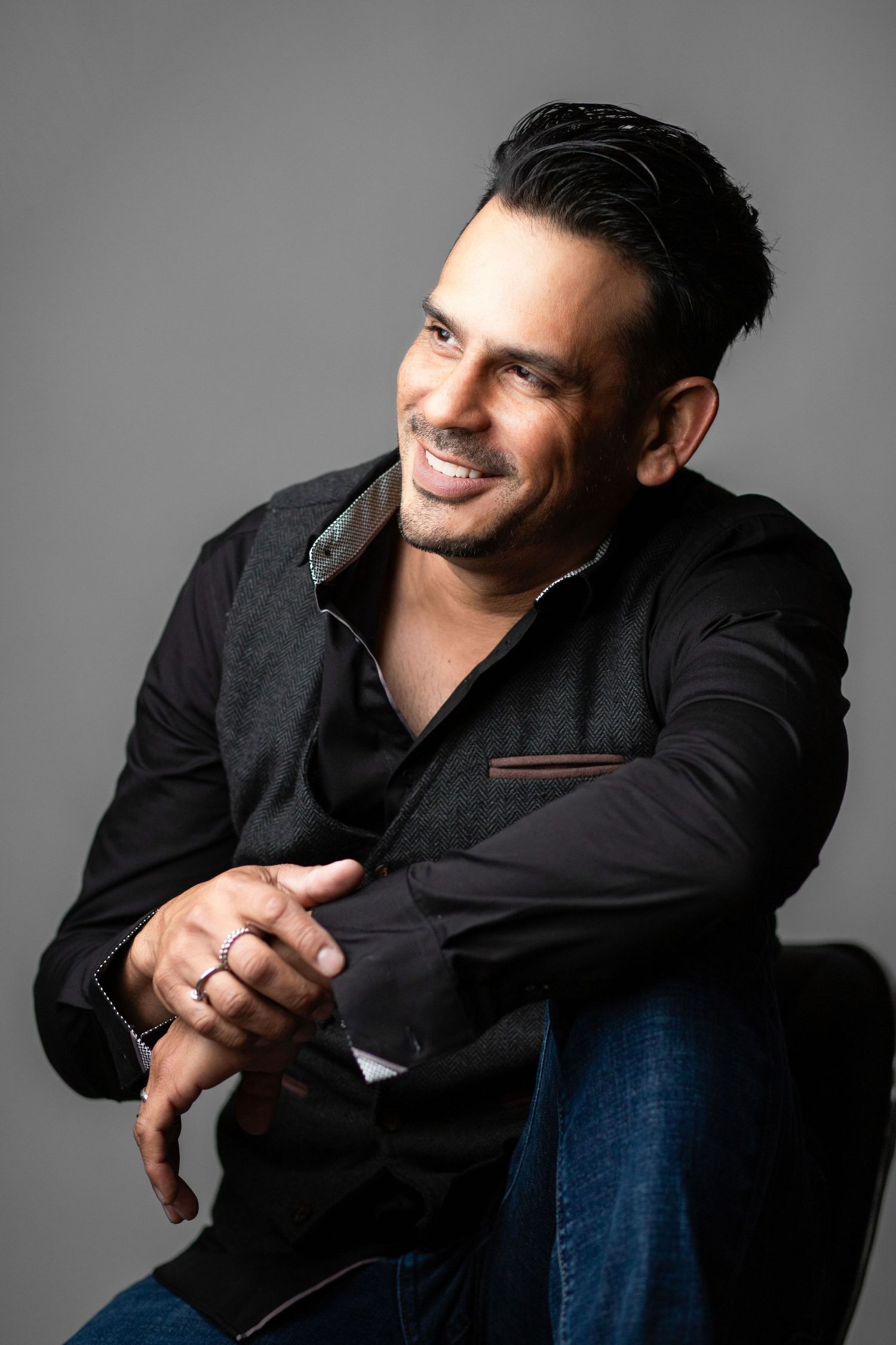 A person in a black shirt and jeans poses against a grey background for a professional portrait.