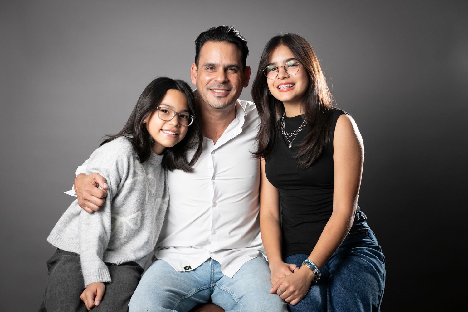 A man and two young girls with glasses smile together in a professional studio portrait against a gray background.