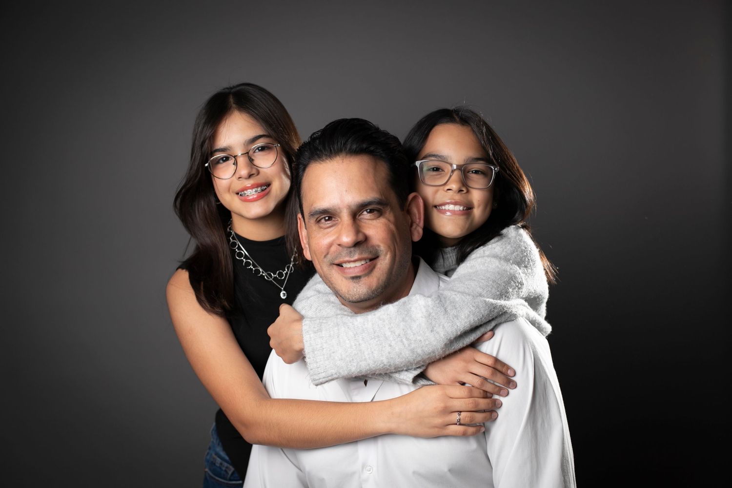 A man smiles with two young girls wearing glasses hugging him in a professional studio portrait.