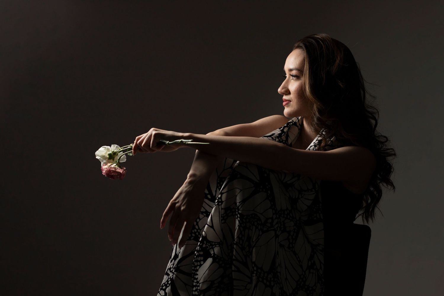 Woman in floral dress sits in chair holding a rose, gazing away in a dark studio setting.