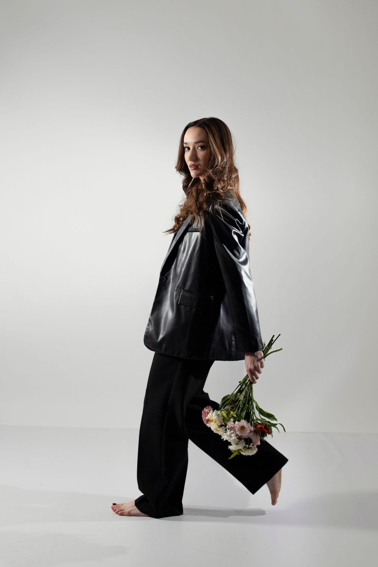 Woman in black leather jacket and trousers holding a bouquet of flowers, posing barefoot against a white background.