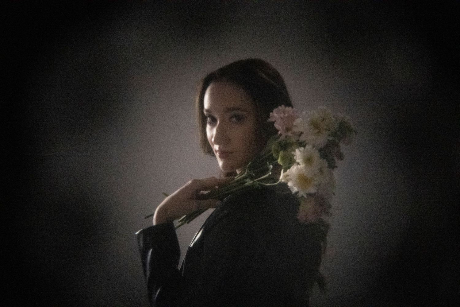 Woman in black jacket holds white and pink flowers over her shoulder against a dark moody background.