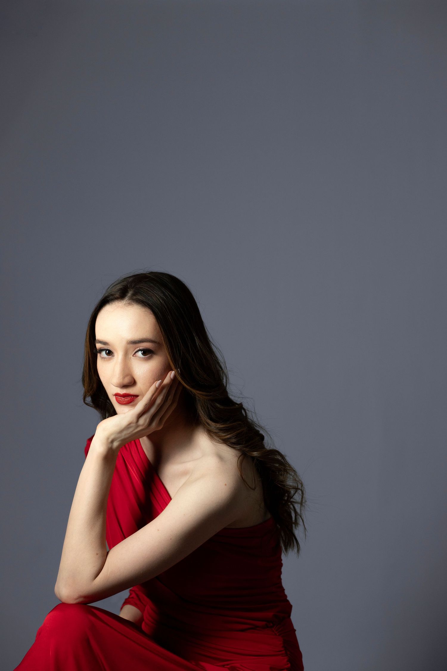 Young woman in red dress with long dark hair poses gracefully against gray background, hand resting on cheek.