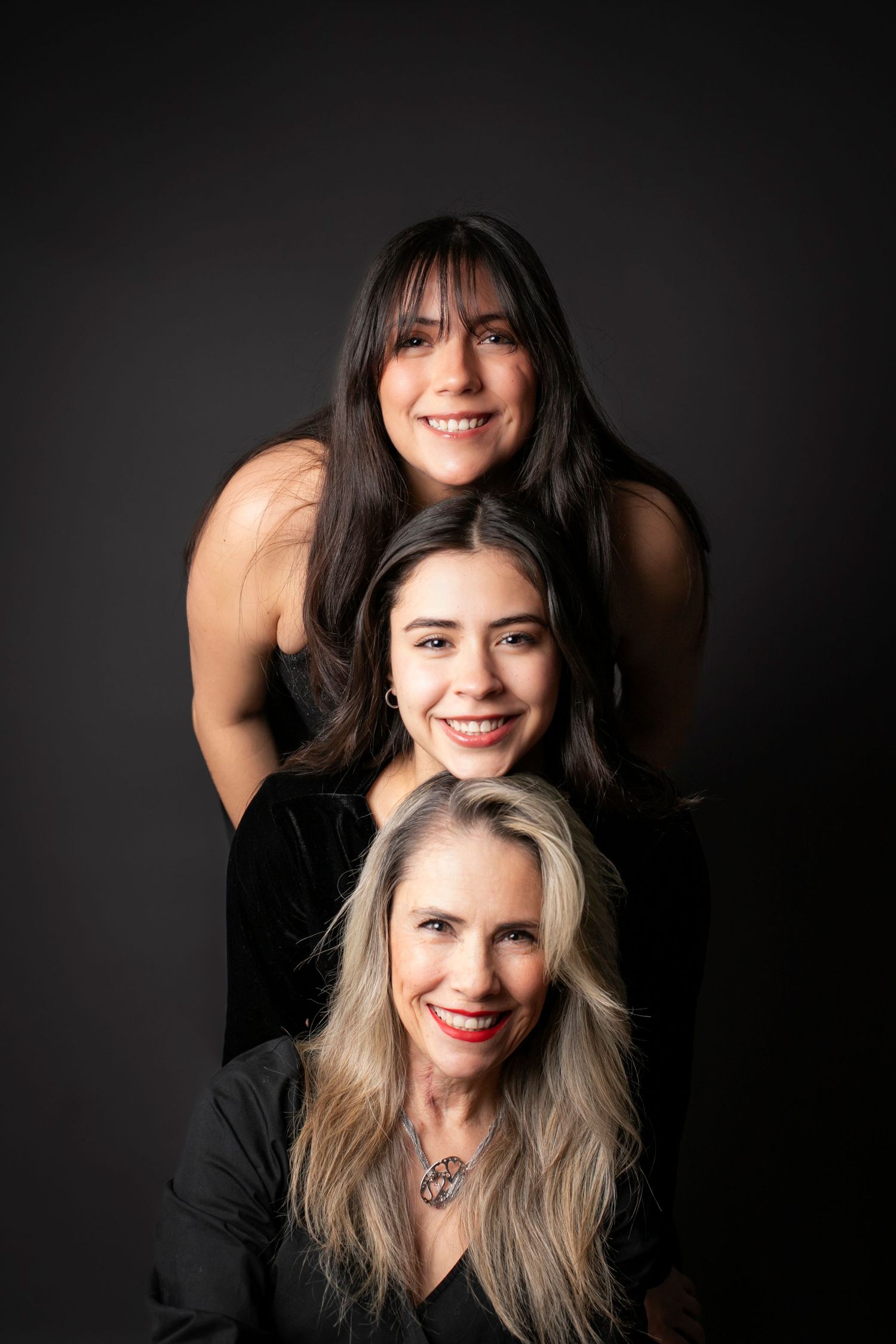 Three smiling women posed in a vertical stack portrait against a dark background, wearing black outfits.