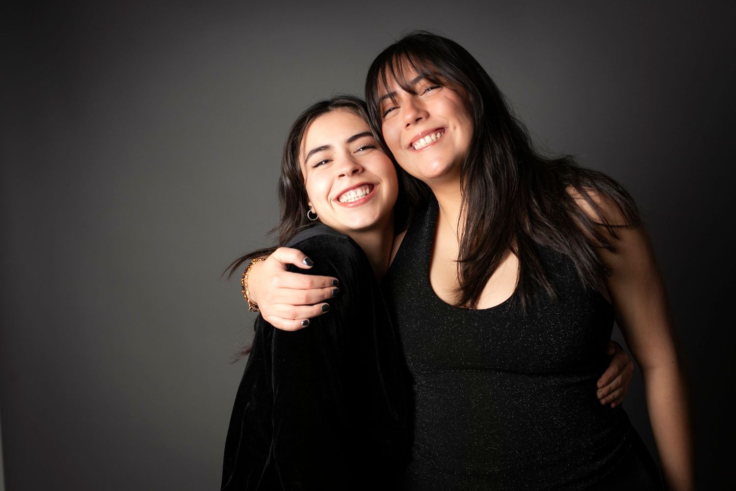 Two young women smiling and hugging each other against a dark gray background.