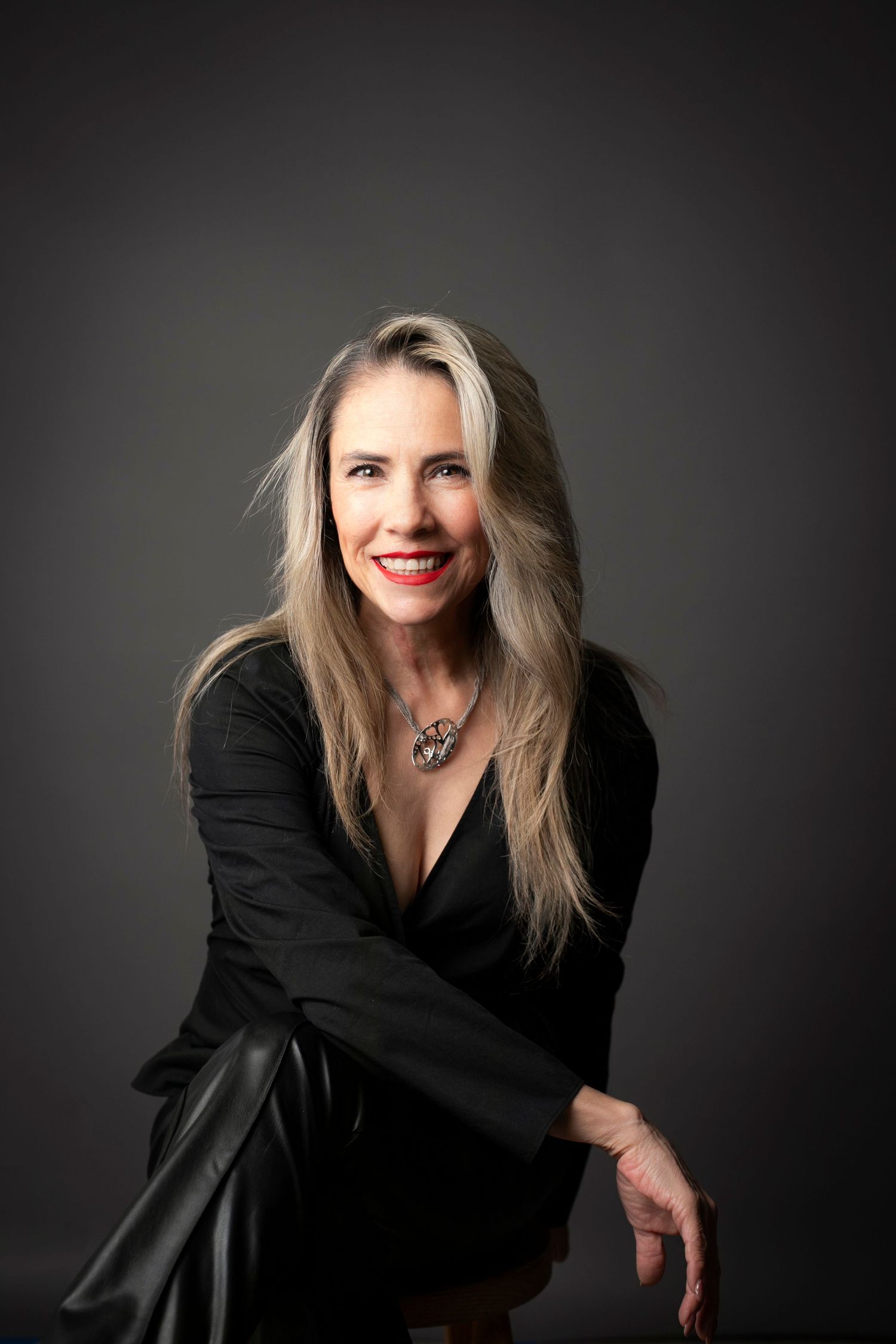 Smiling woman with long blonde hair wearing a black dress and silver necklace poses confidently in a studio setting.