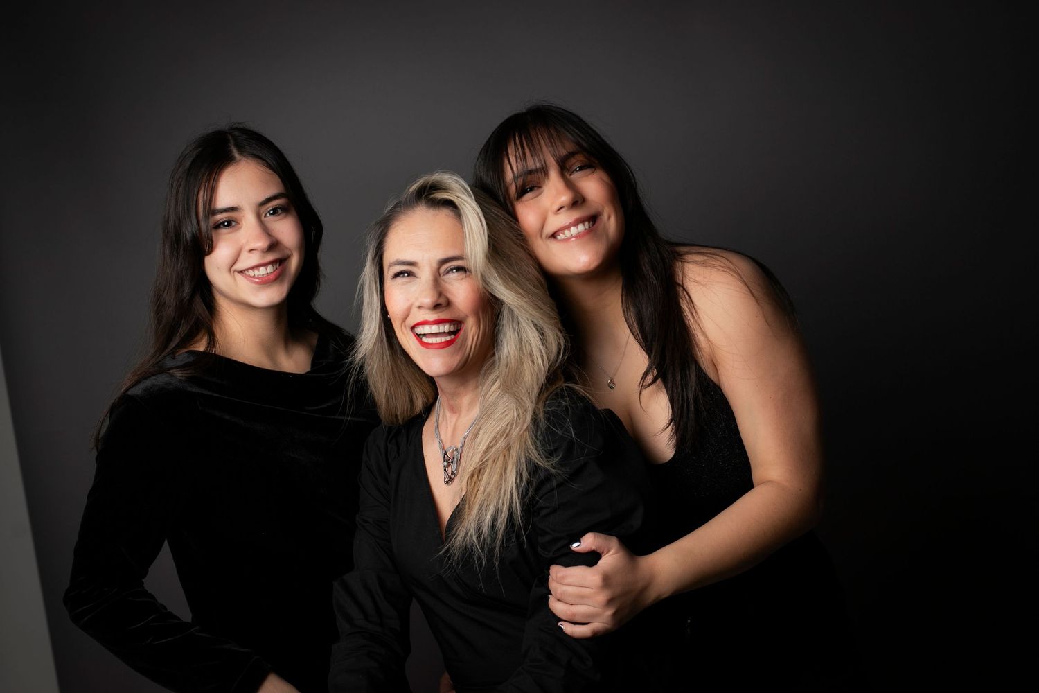 Three women in black outfits smiling together against a dark gray background in a professional portrait photo.