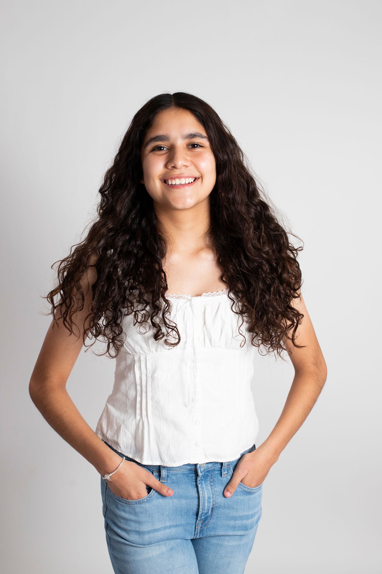Smiling teenage girl with long curly hair wearing a white sleeveless top and jeans, hands in pockets.