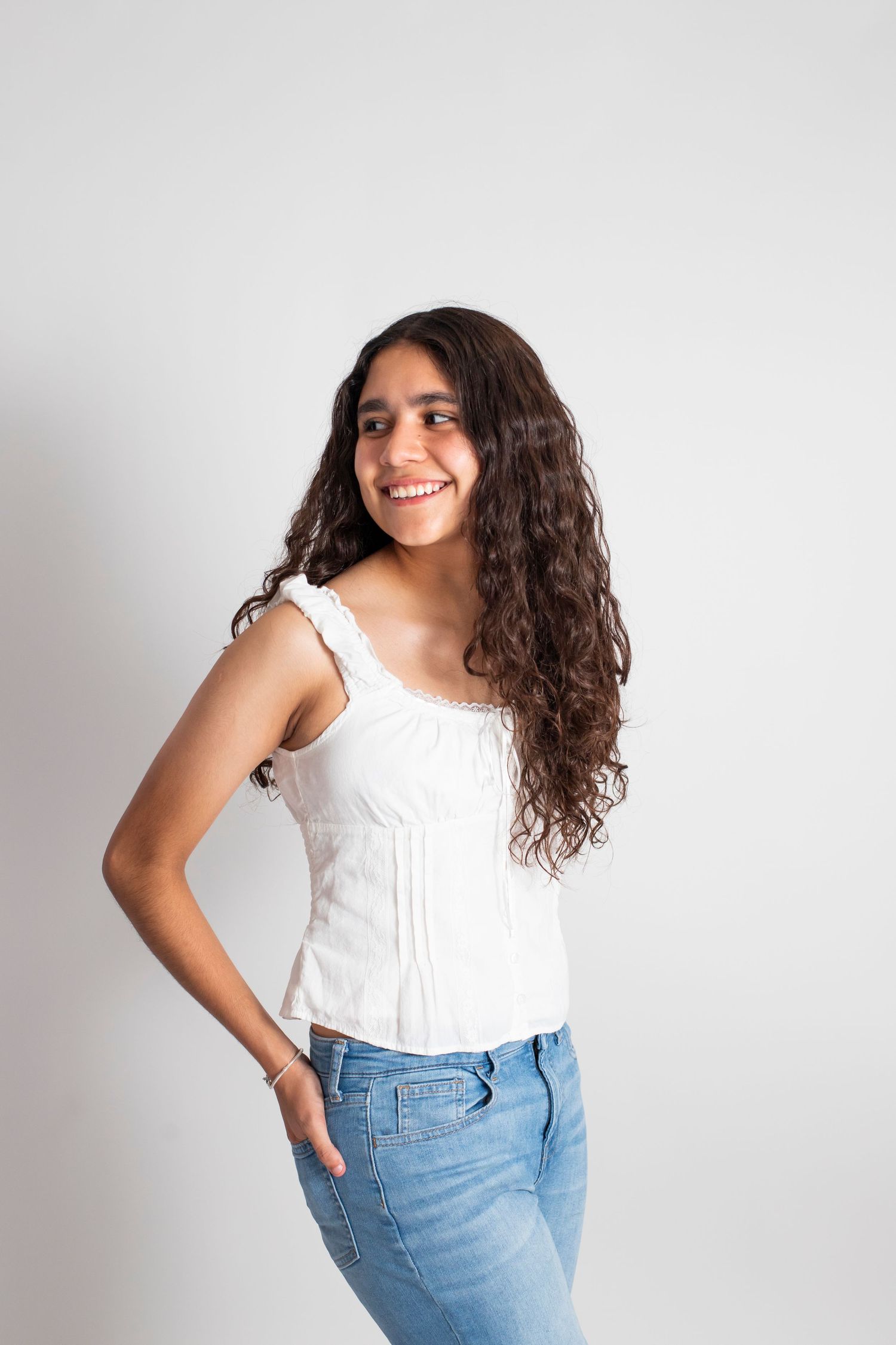 Smiling young woman with curly hair wearing a white top and jeans poses against a light gray background.