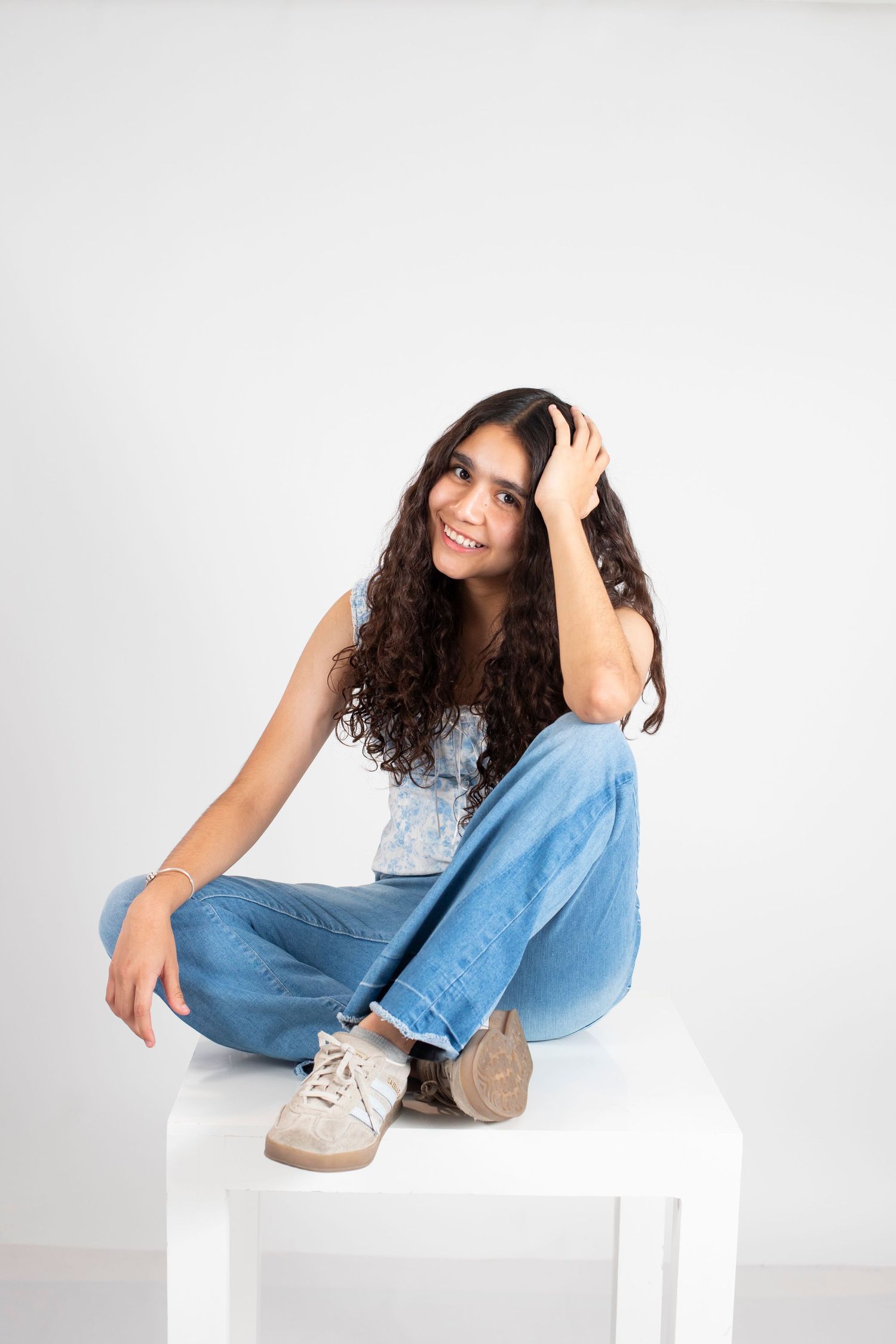 Young woman with curly hair sitting cross-legged on white cube, wearing blue jeans and tank top, smiling confidently.