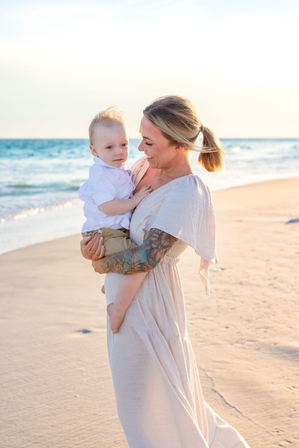A woman in a flowing white dress holds a small child while standing on a sunny beach at sunset with ocean waves behind them.