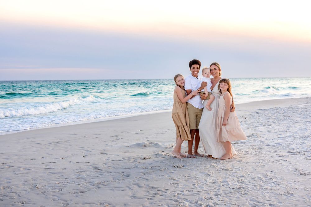 A group embraces on a white sandy beach at sunset with turquoise waves and pink sky in the background.
