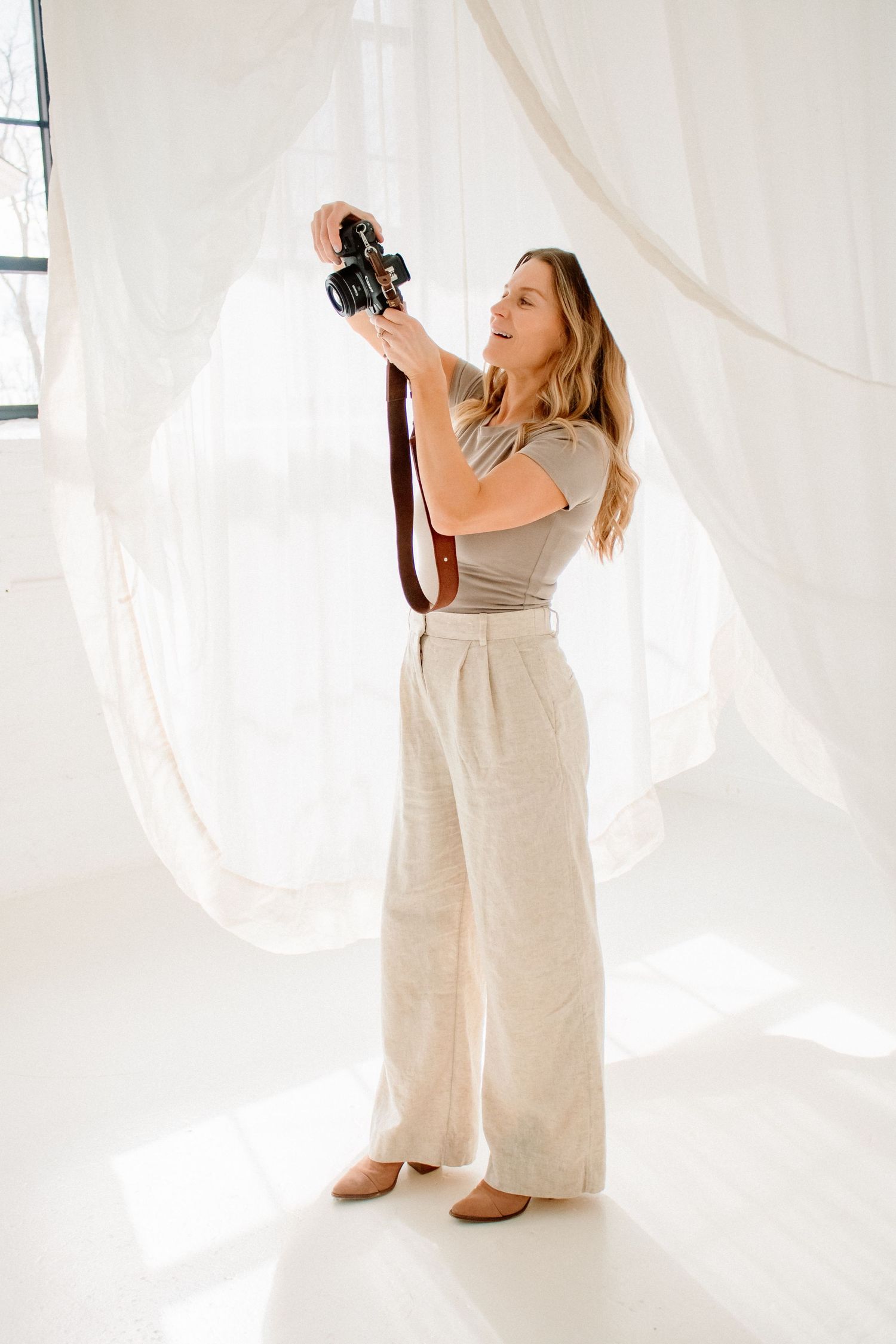 Professional photographer in beige linen outfit shooting with vintage camera against white flowing backdrop.