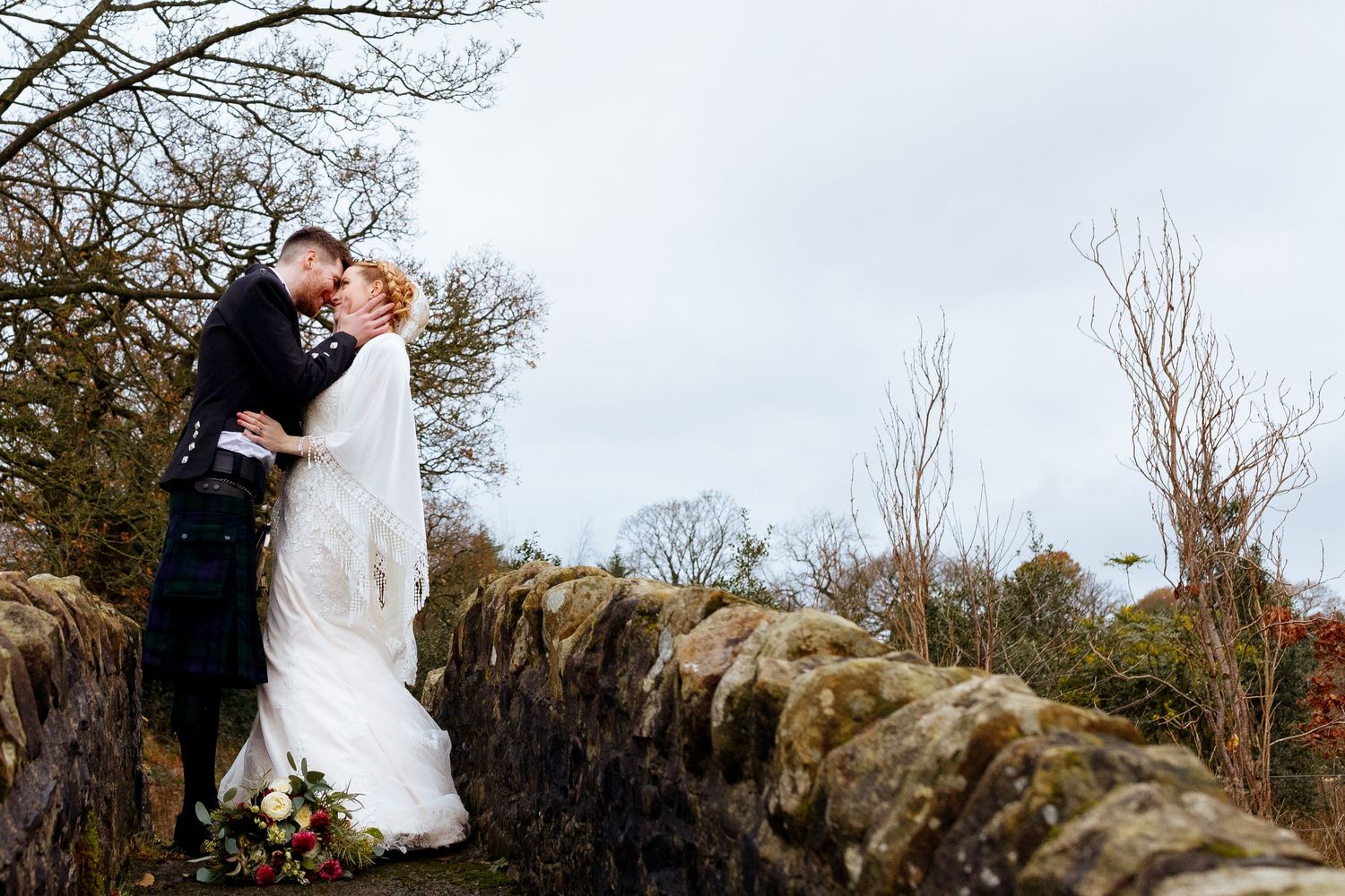 A couple shares a romantic kiss atop a stone wall surrounded by autumn trees beneath an overcast sky. The Outbarn, Clitheroe.
