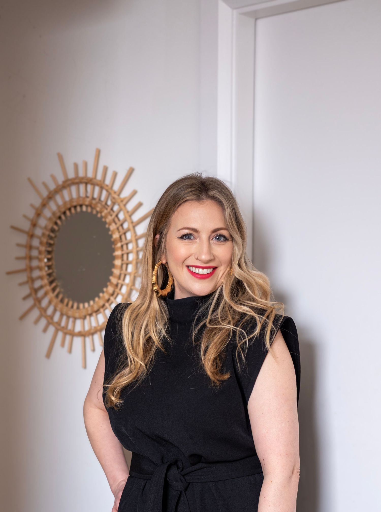 Woman in black dress poses next to decorative sunburst mirror on white wall.