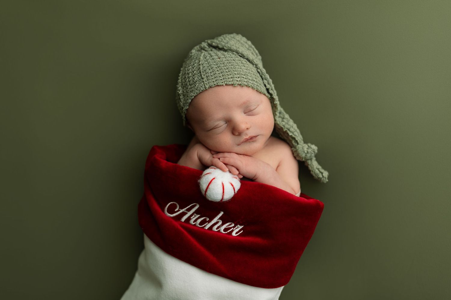 Newborn photography session with green knit hat and red blanket against dark green backdrop.