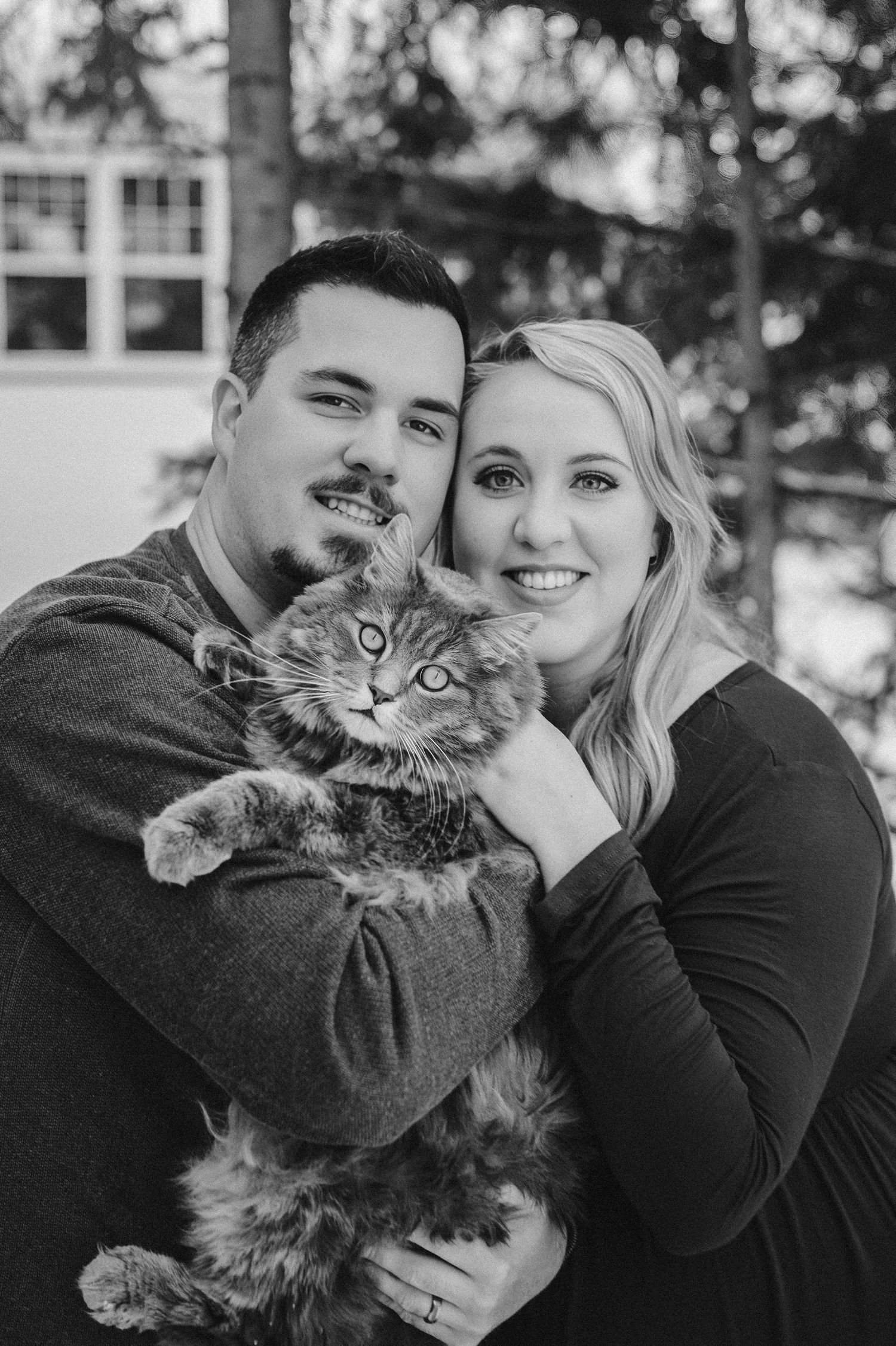 A couple holds their fluffy grey cat in a black and white portrait.