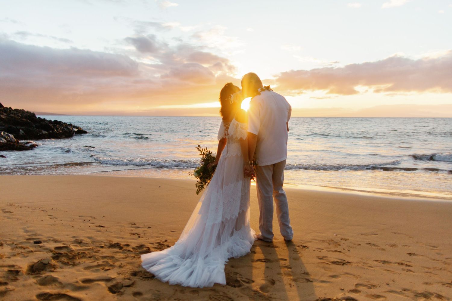 A couple in wedding attire embrace on a sandy beach at sunset with ocean waves in the background.