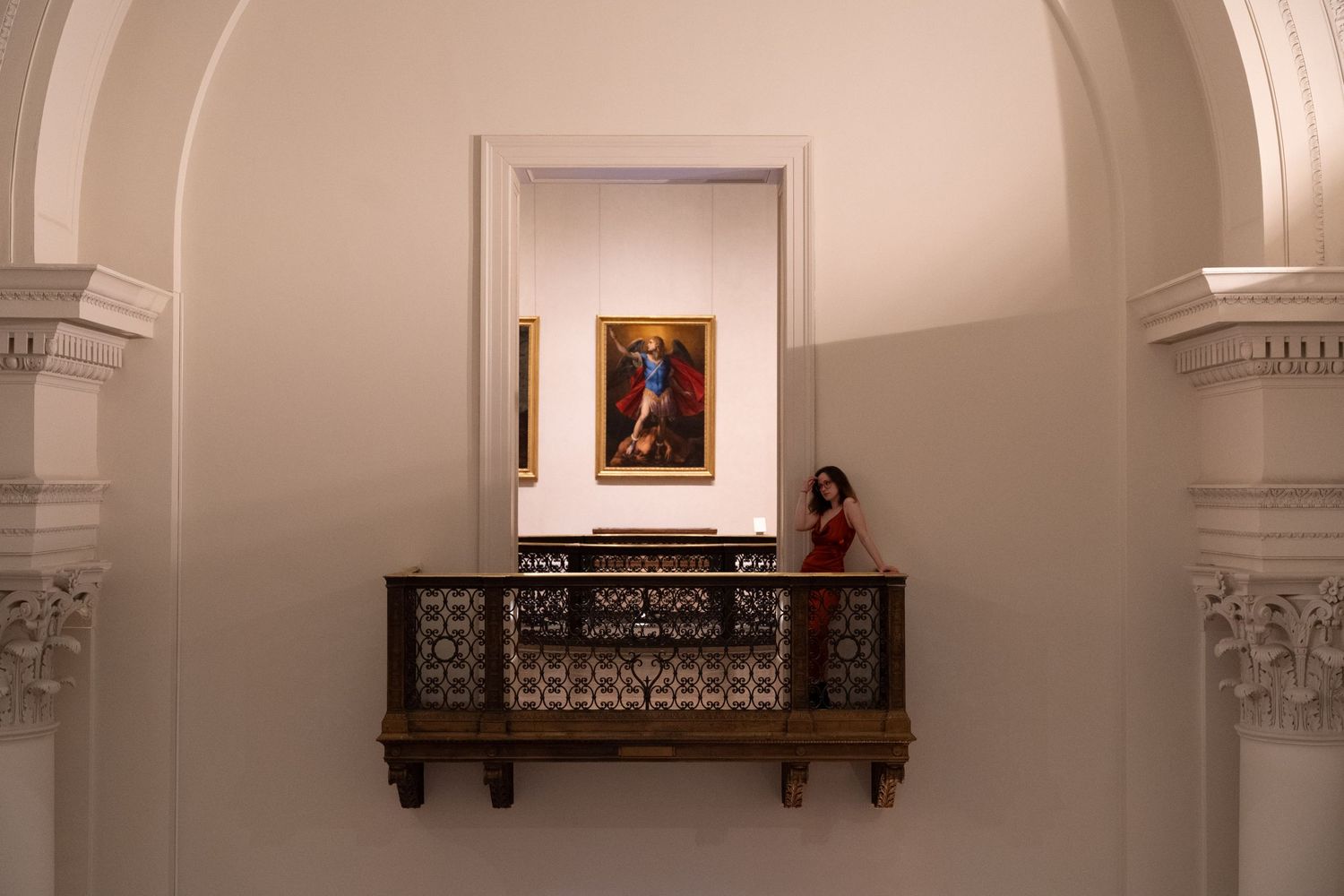 A framed portrait painting hangs above an ornate balcony railing inside an elegant arched hallway.