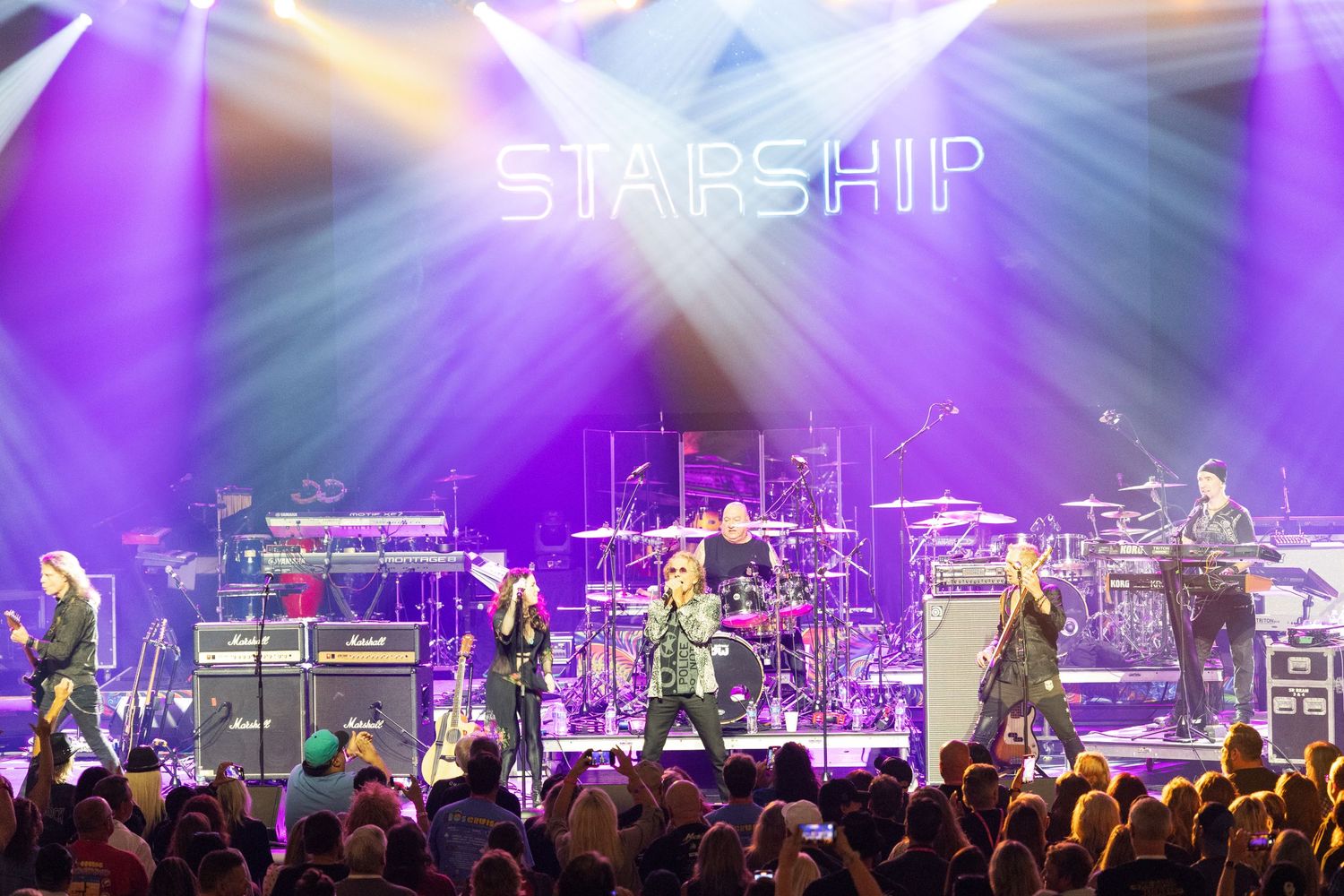 A rock band performs on stage with bright purple lighting and 'STARSHIP' text illuminated above the crowd.