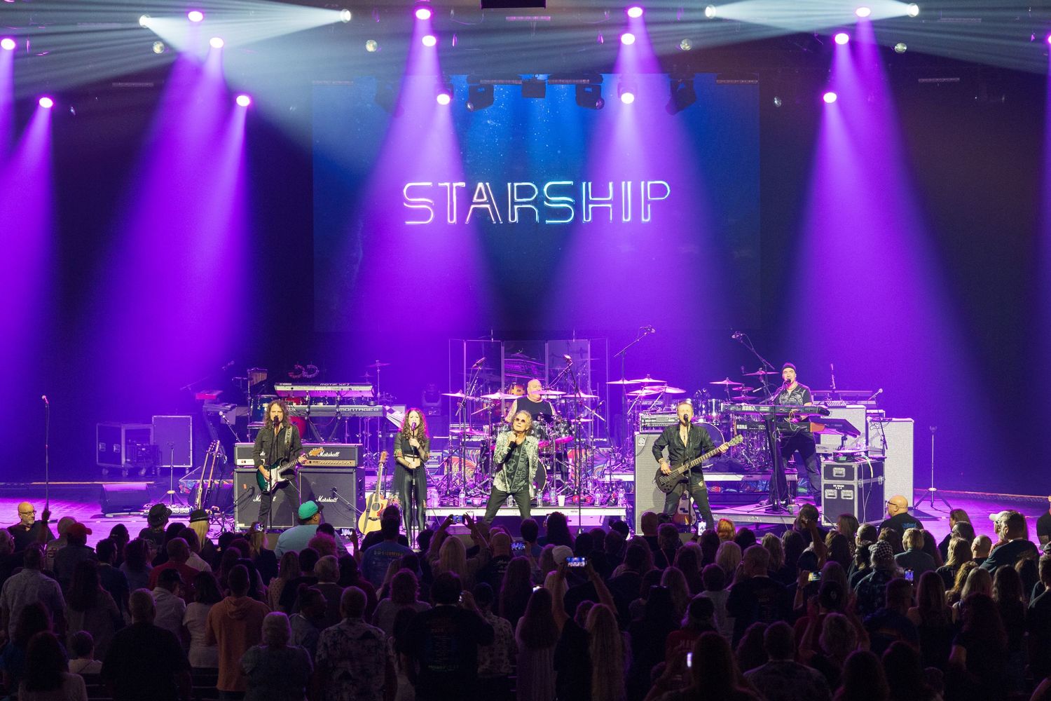 Rock band Starship performs live on stage with purple and blue concert lighting illuminating their musical equipment and crowd.