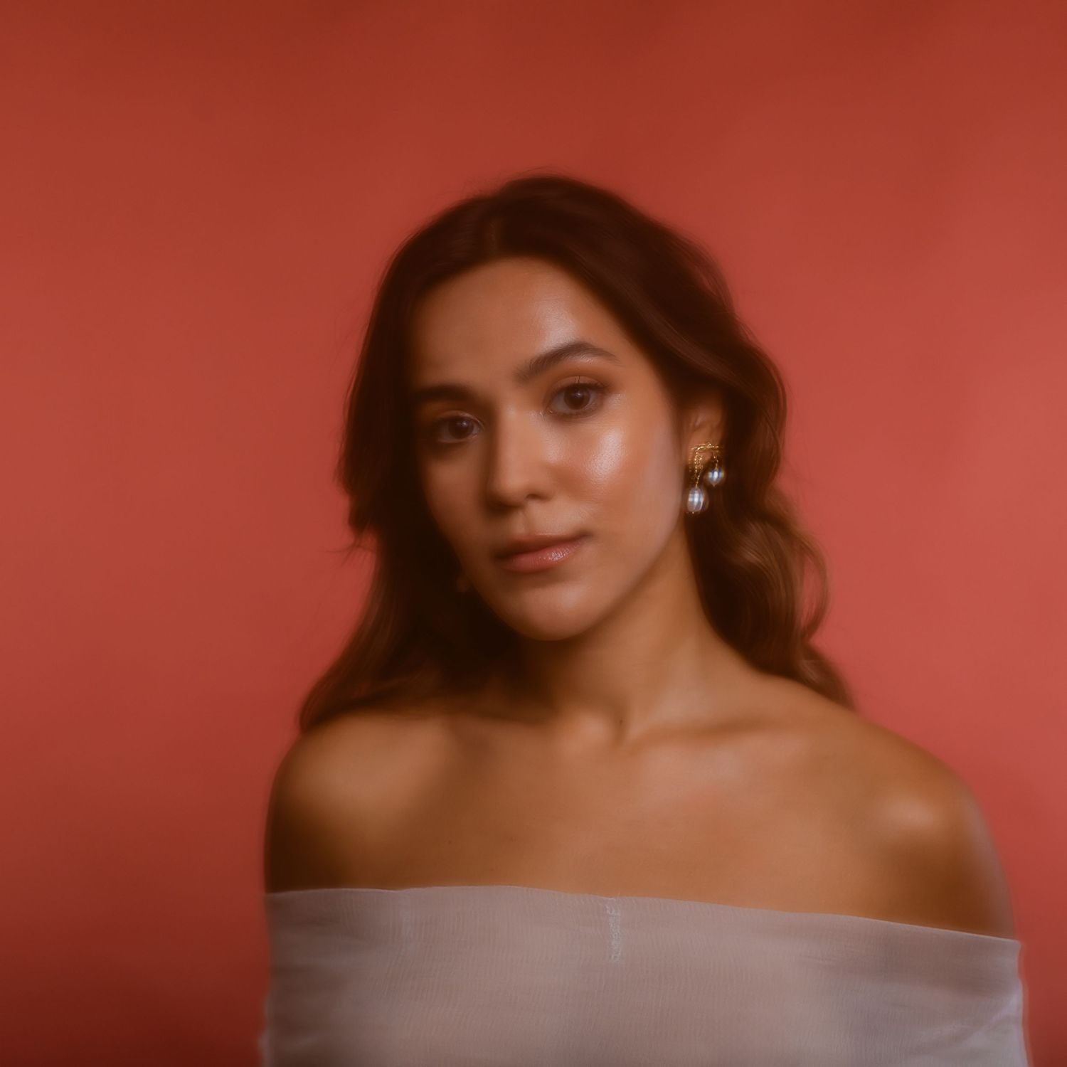 Portrait with soft lighting against terracotta background shows bare shoulders in mellow tones with gold earrings.
