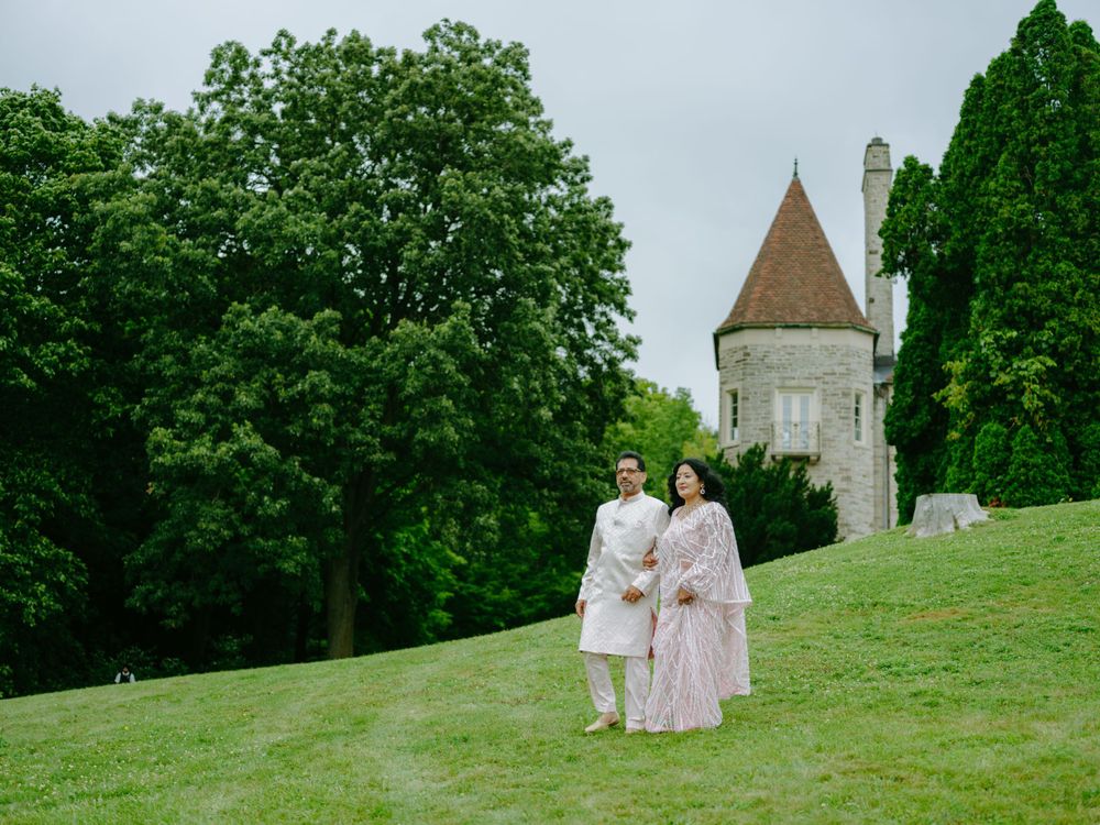 A couple in elegant attire stands on a lush green lawn with a medieval castle tower in the background.