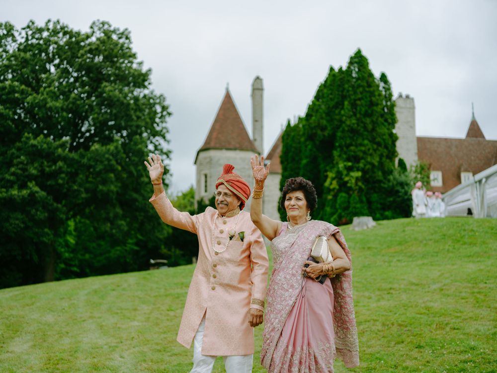 A couple in traditional Indian wedding attire stand together on a grassy lawn with a castle-like building in the background.