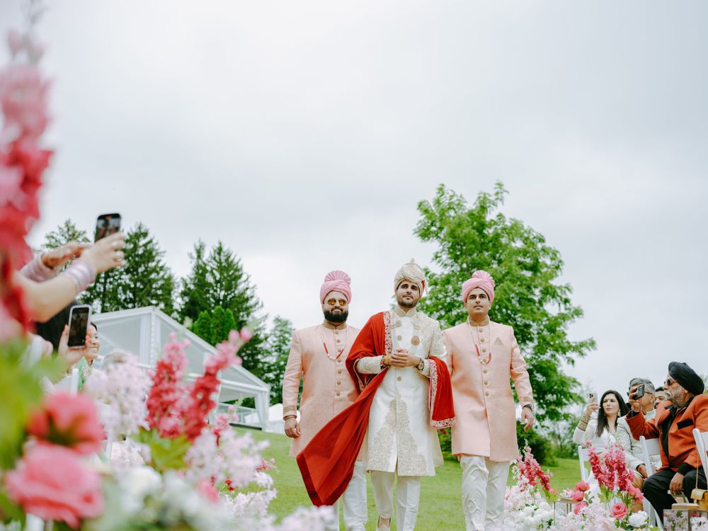 Joyful wedding celebration outdoors with people in traditional Indian attire surrounded by pink and red flowers.
