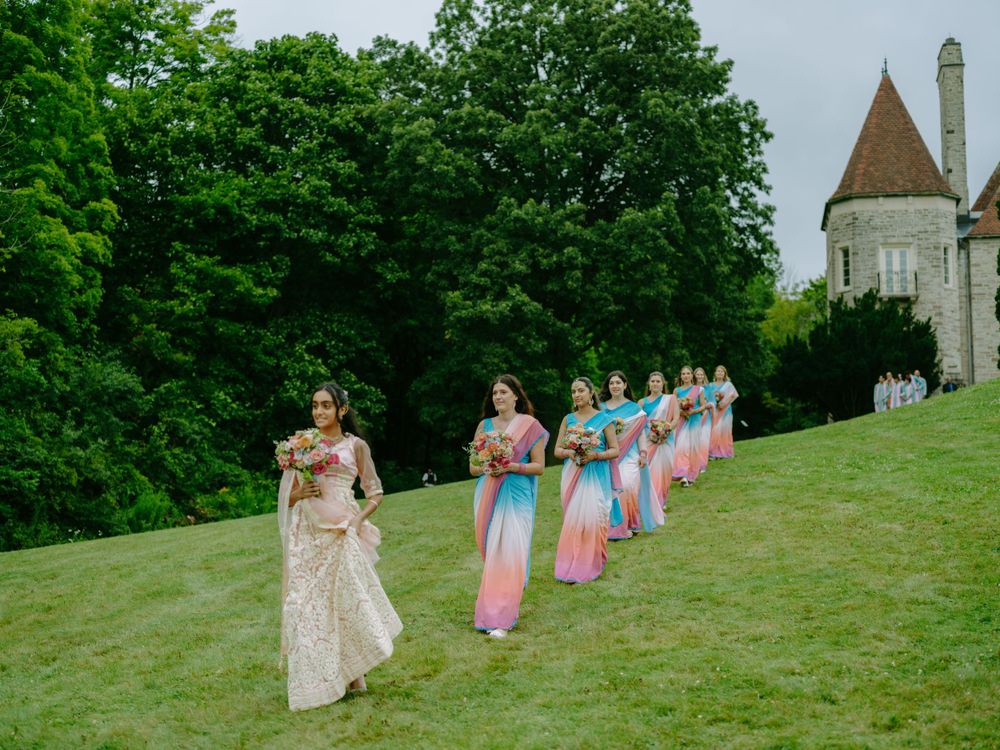 Wedding party in pink dresses walks in a line on a grassy hill with a historic stone tower and trees in the background.