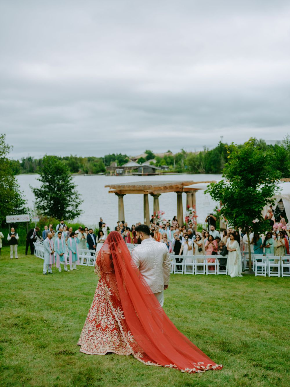 A couple in traditional wedding attire shares a moment under a lakeside gazebo with guests watching from white chairs.