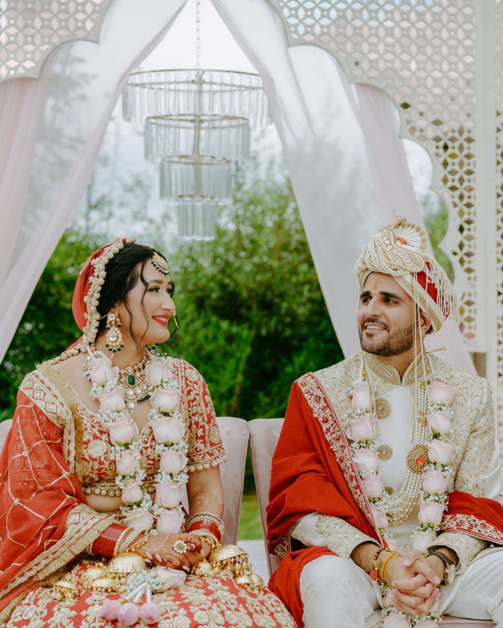 Traditional Indian wedding ceremony taking place outdoors with couple in ornate red and white attire under a decorated mandap.