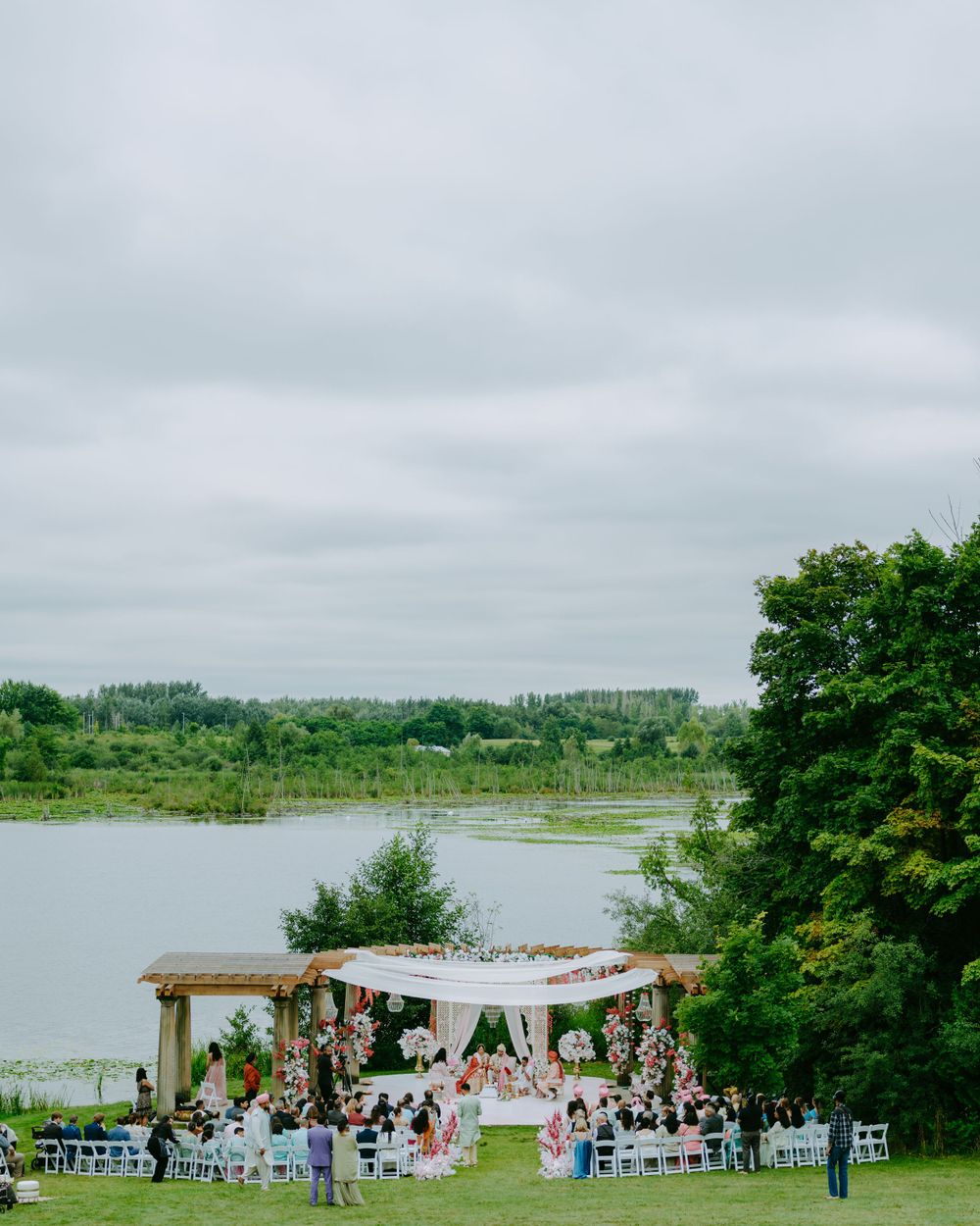 An outdoor wedding ceremony setup with a wooden pergola and floral decorations by a serene lake under cloudy skies.