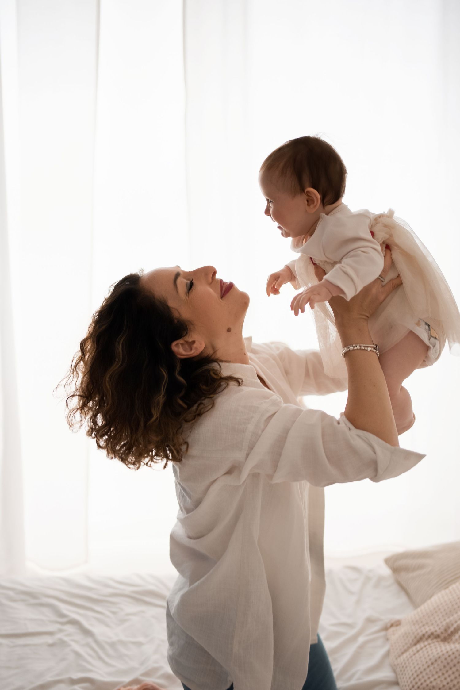 A curly-haired mother joyfully lifts her smiling baby in a bright, airy bedroom.