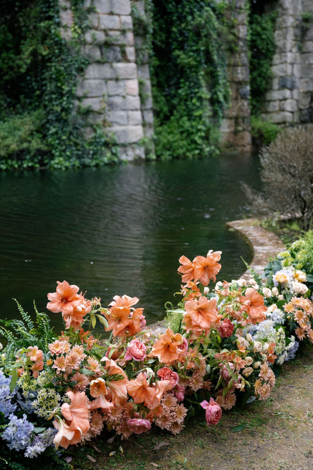 Garden flowers in peach and coral tones border a tranquil pond by stone walls with trailing ivy.