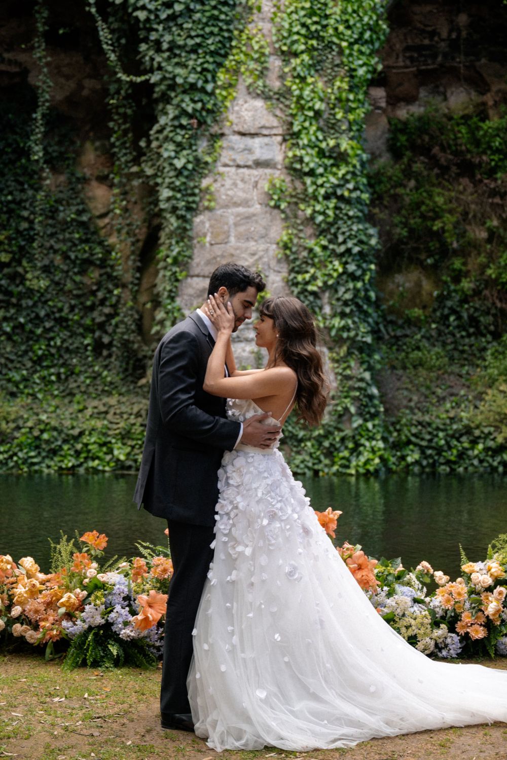 Romantic wedding photo by ivy-covered stone wall and pond with peach floral arrangements.