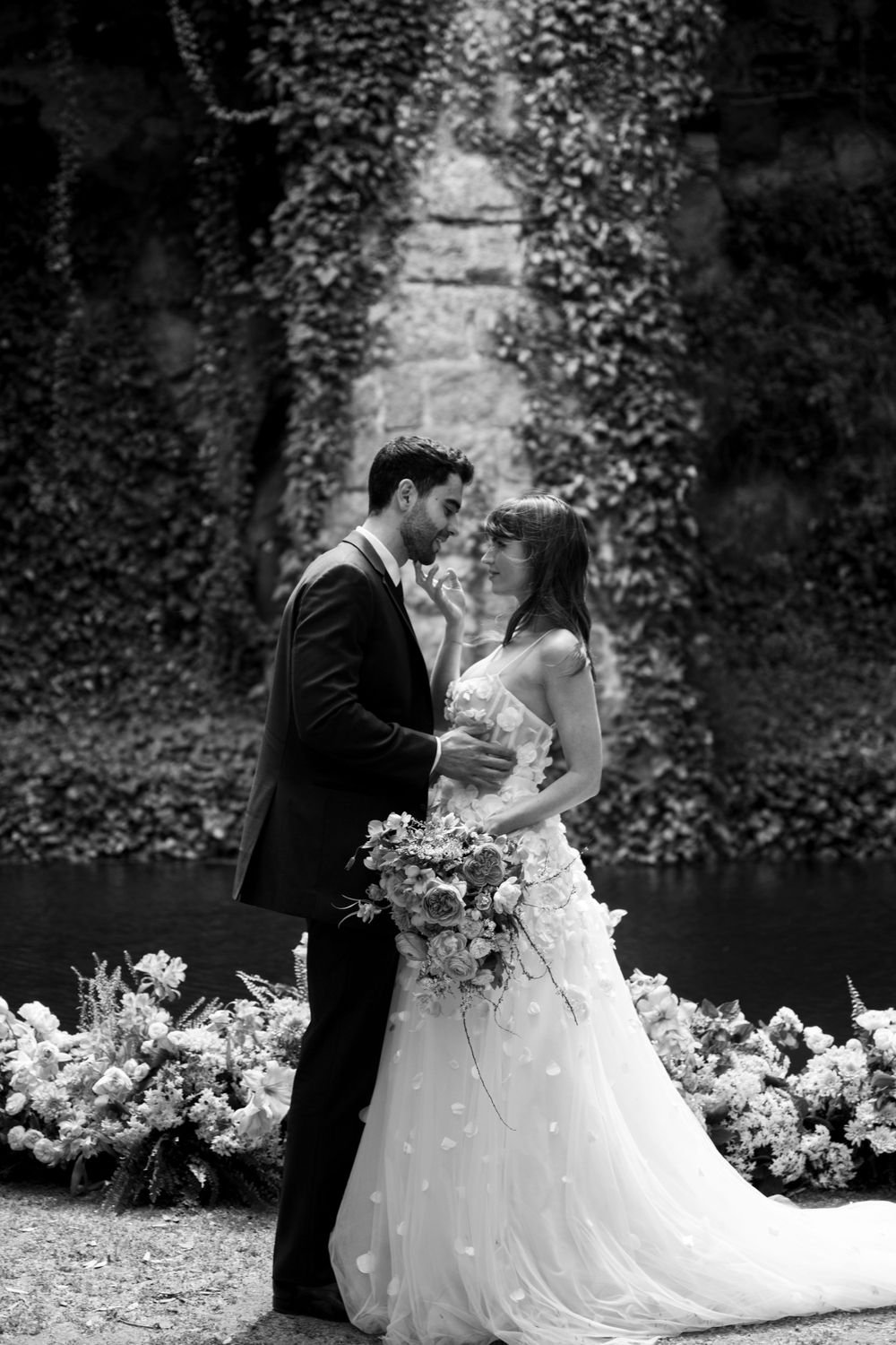 Black and white wedding portrait against ivy-covered stone wall with floral decor.