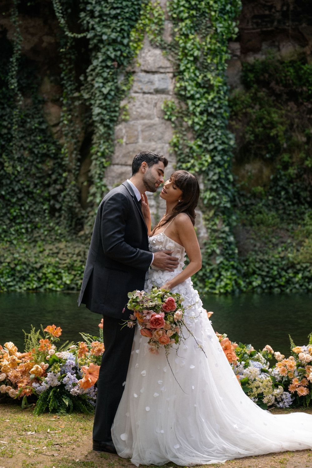 Romantic wedding portrait with couple surrounded by peach flowers and ivy-covered stone wall.
