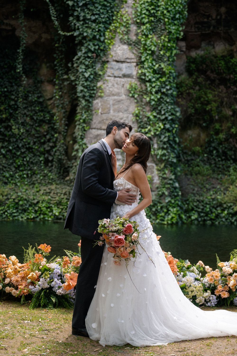A romantic wedding ceremony by a pond with ivy-covered walls and peach floral arrangements.