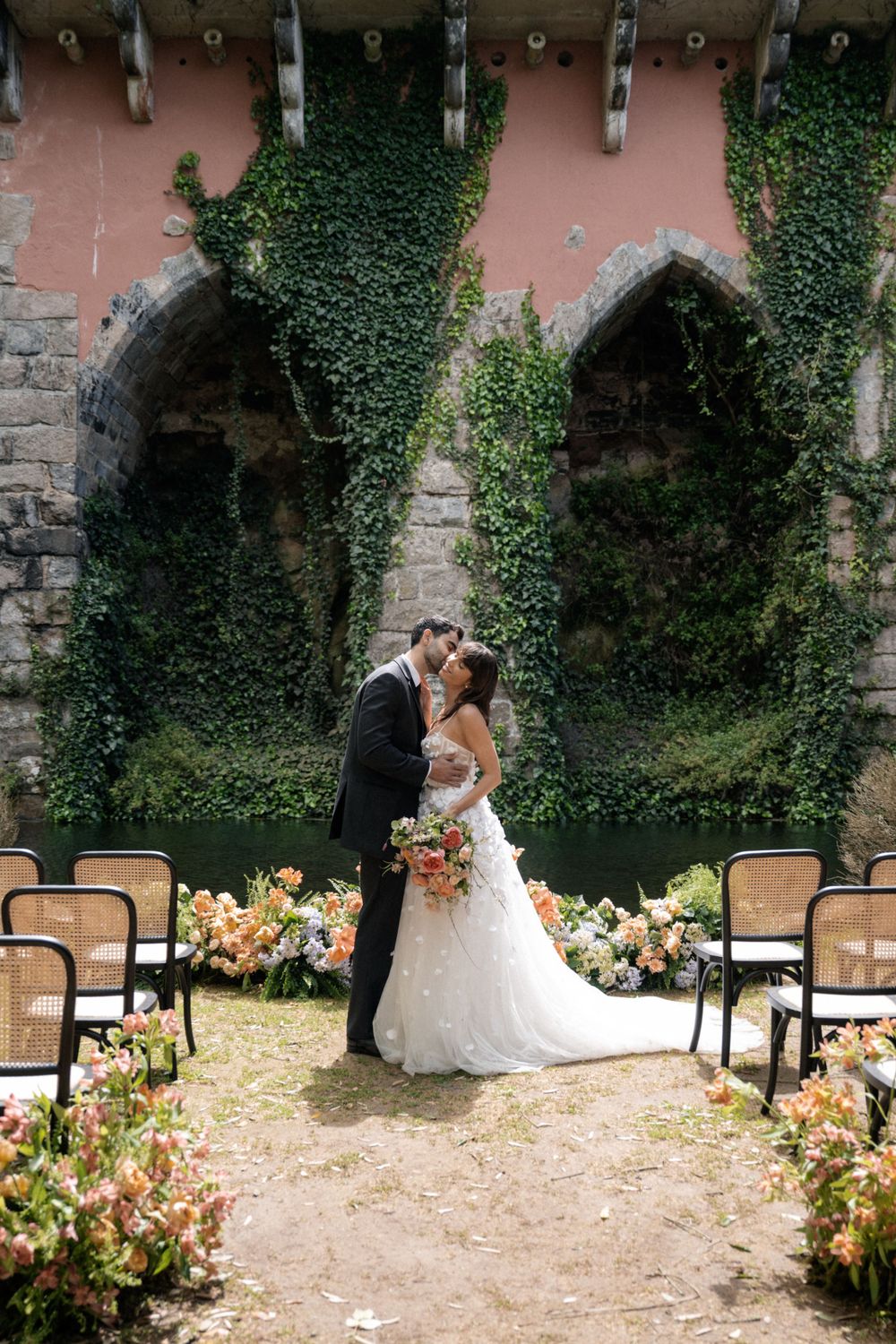A wedding ceremony setup with cane chairs and peach floral arrangements against stone arches covered in ivy.