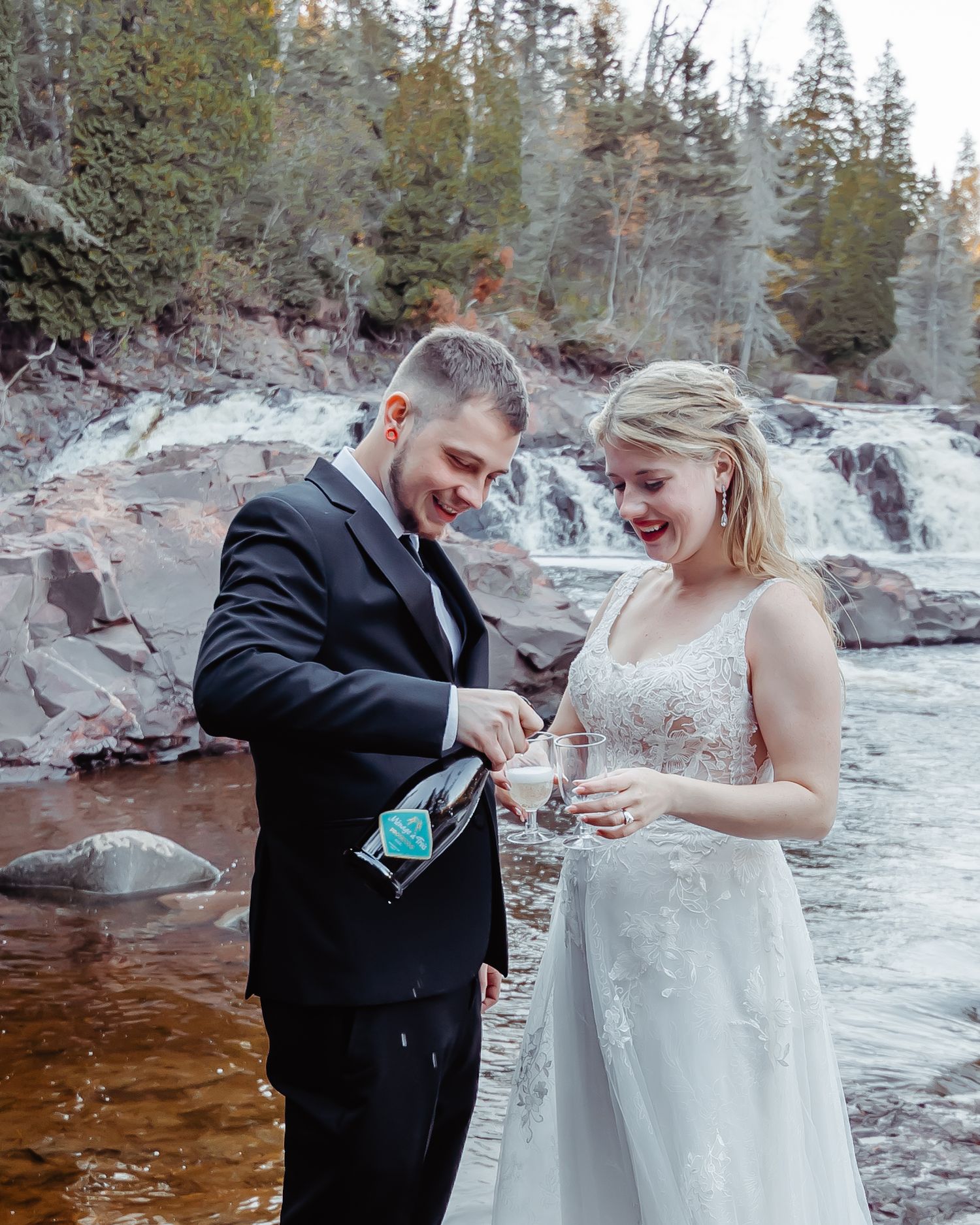 A couple in wedding attire shares a joyful moment pouring drinks by a scenic waterfall in a forest setting.